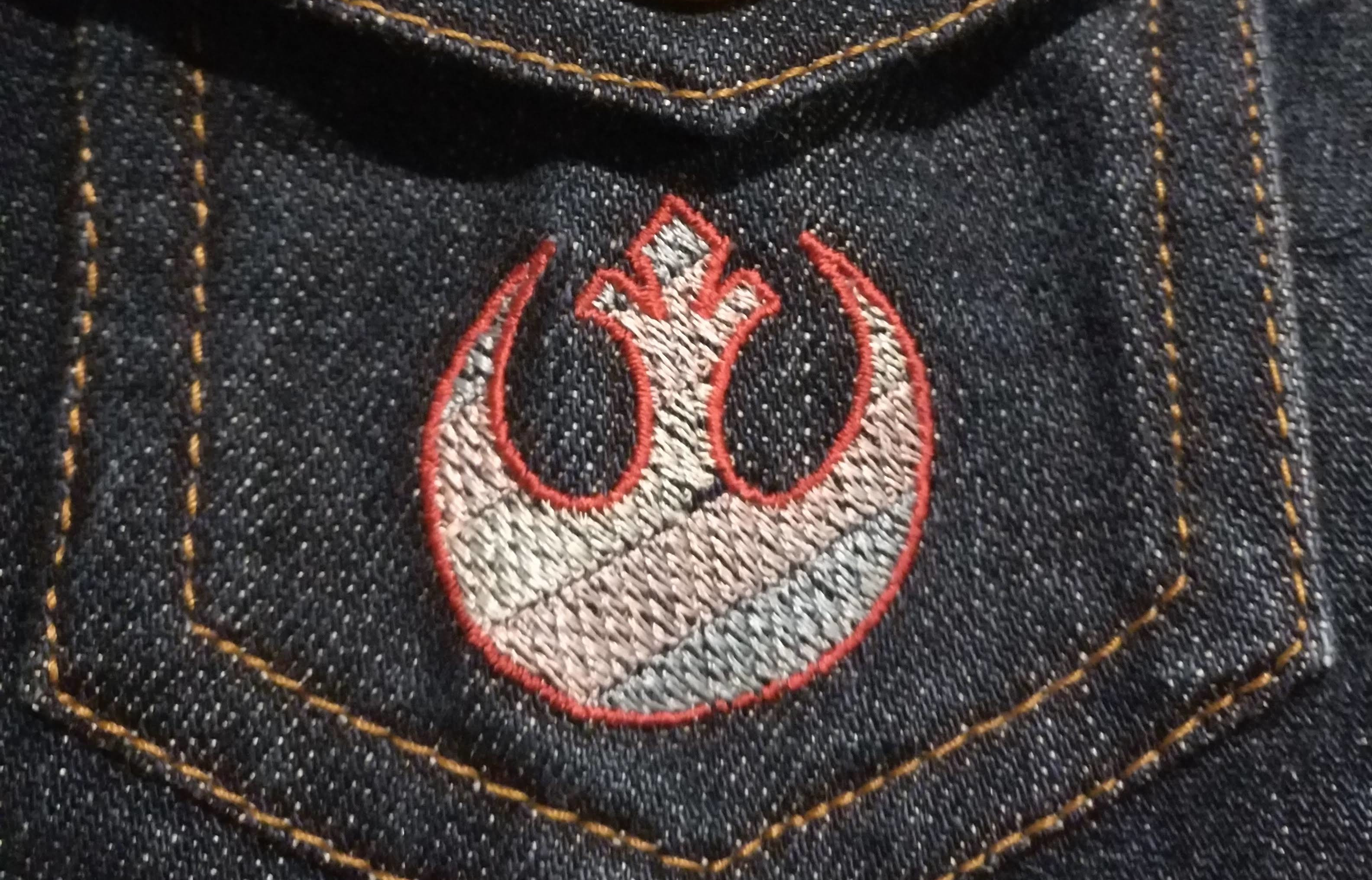 An embroidered Trans pride flag in the shape of the Star Wars Rebel Alliance logo